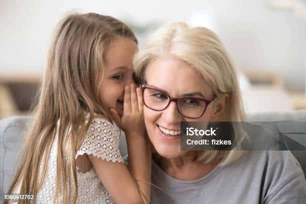 Cute little granddaughter whispering in ear telling secret to understanding smiling grandma, kid girl secretly talking to granny having fun gossiping, trust in grandmother and grandchild relations